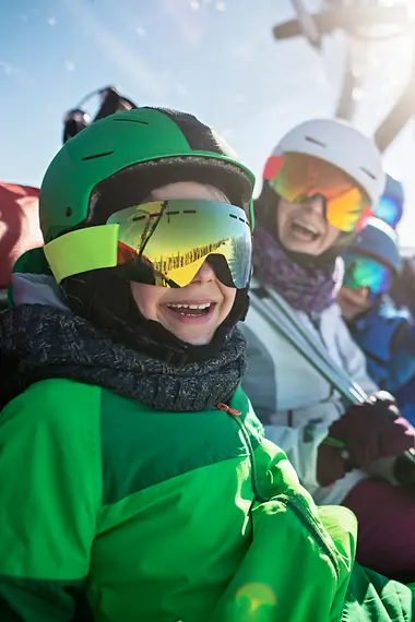 Wintersport reisverzekering Een gezin zit in een skilift en kijkt lachend uit voor de camera.
